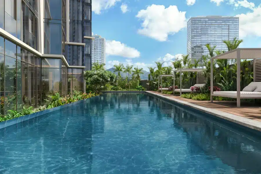Rectangular outdoor swimming pool beside a modern glass building, surrounded by palm trees, cabanas, and city high-rises under a partly cloudy sky.