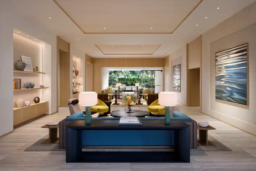Modern living room with neutral tones, blue sofa, two table lamps, decorative shelves, large wall art, and a view of a garden through glass doors in the background.