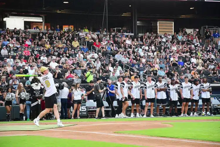 A baseball player swings at a pitch during a game, while teammates watch from the sidelines and a large crowd fills the stadium seats.