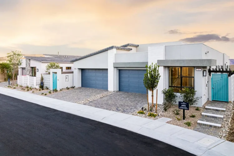 Modern duplex with flat roof, two blue garage doors, and minimal landscaping, located on a clean, paved street at sunset. A for-sale sign is posted near the entrance.