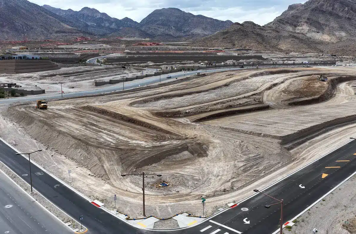 Aerial view of a large construction site with dirt roads and heavy equipment near a highway, surrounded by mountains and residential areas in the background.