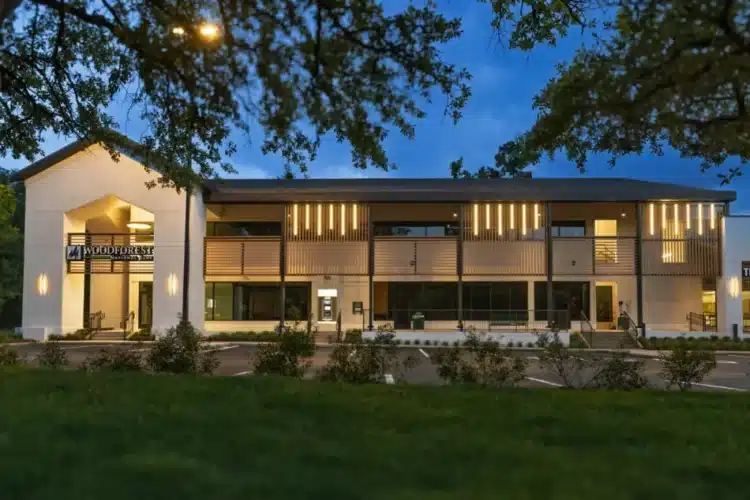 A two-story modern building with large windows and vertical lights on the upper facade, viewed at dusk with trees and grass in the foreground.