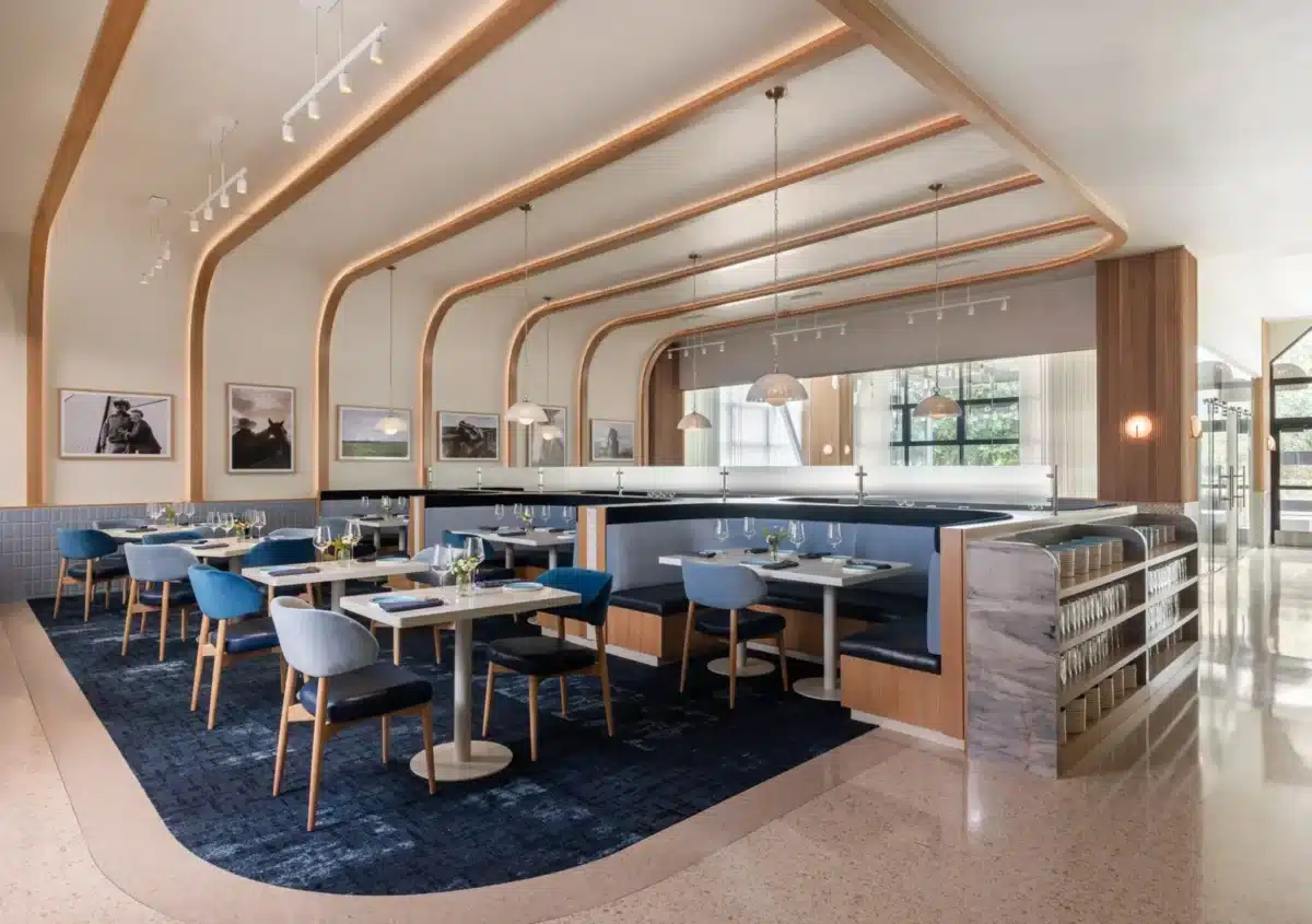 Modern restaurant interior with blue chairs, booth seating, set tables, contemporary lighting, wall art, and large windows letting in natural light.