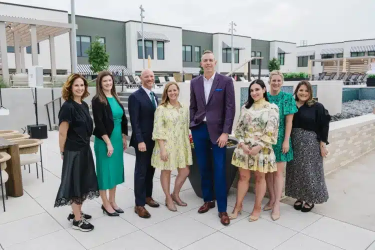 Nine people, dressed in business or semi-formal attire, stand in a row outdoors on a patio with modern buildings in the background.
