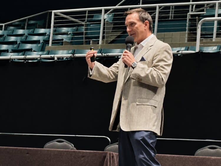 Man in a beige blazer holding a microphone speaks on stage in front of empty green seats, gesturing with his other hand.