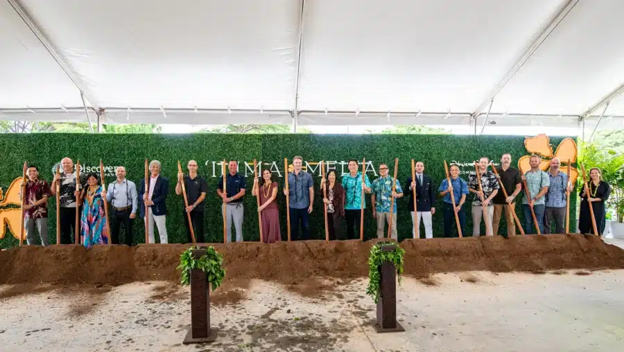 A group of people stands in a row holding shovels at a groundbreaking ceremony in front of a green backdrop under a large tent.