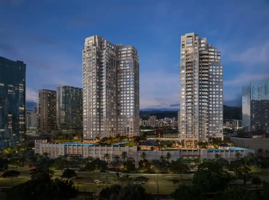 Two modern high-rise residential towers are illuminated at dusk, surrounded by other buildings. Trees line the foreground, with mountains visible in the background.