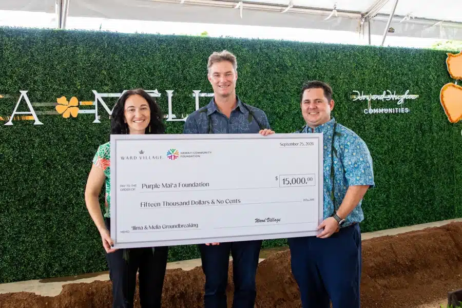 Three people stand smiling and holding a large ceremonial check for $15,000 made out to the Purple Mai‘a Foundation at an event.