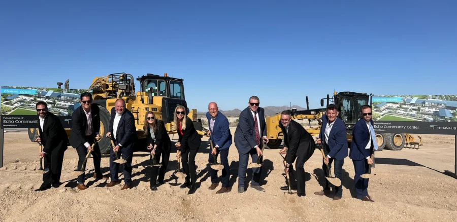 Ten people in business attire break ground with shovels at a construction site, with large construction vehicles and project banners in the background under a clear blue sky.
