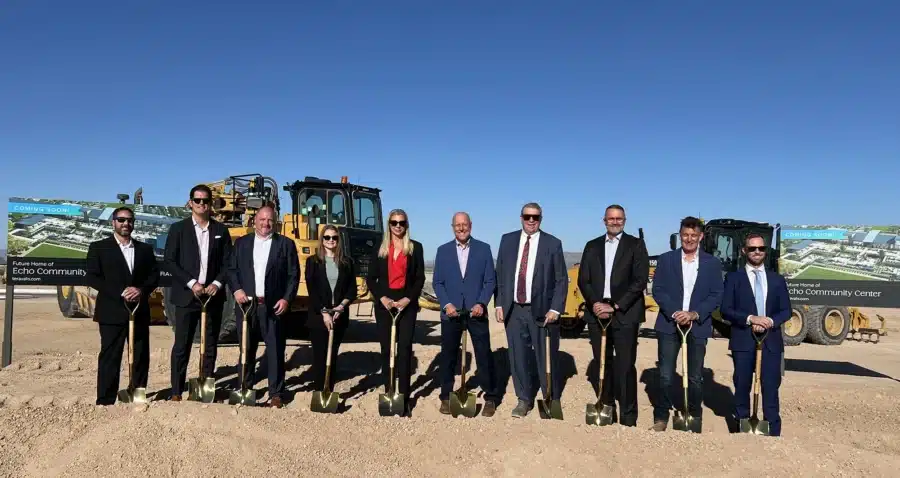 Nine people in business attire stand in a row with shovels at a construction site, ready for a groundbreaking ceremony. Construction vehicles and project signage are visible in the background.
