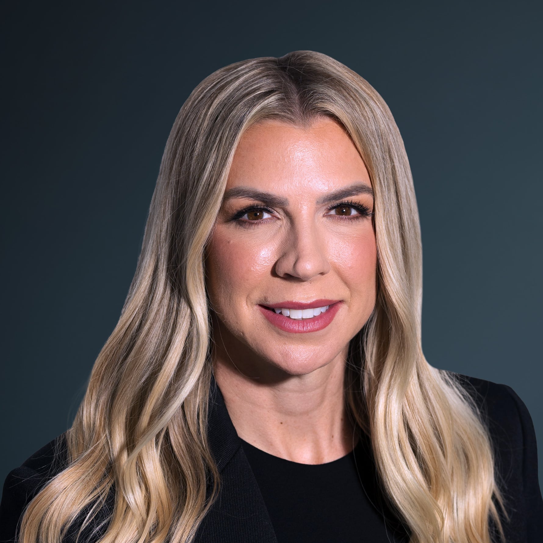 Woman with long blonde hair, wearing a black blazer and top, smiling slightly, posed against a dark background.