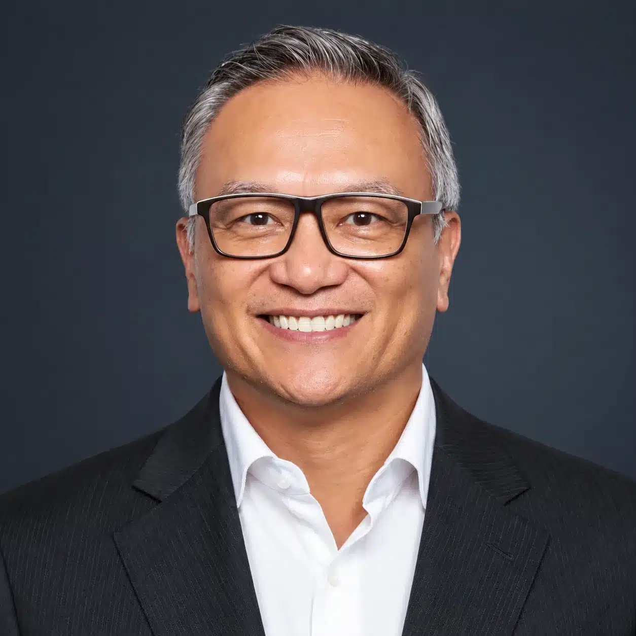 Smiling man with short gray hair and glasses, wearing a dark suit jacket and white collared shirt, posed in front of a plain dark background.