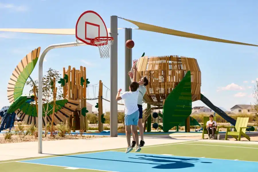 Two people play basketball on an outdoor court near modern playground equipment on a sunny day.