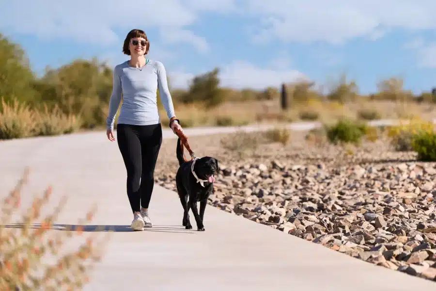 Woman wearing sunglasses walks a black dog on a paved path through a park with rocks and desert plants under a blue sky.