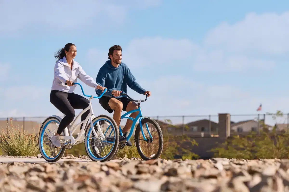 Two people ride bicycles side by side on a rocky path, with a fence and buildings in the background under a blue sky.