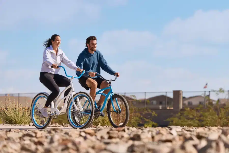 Two people ride bicycles side by side on a rocky path, with a fence and buildings in the background under a blue sky.