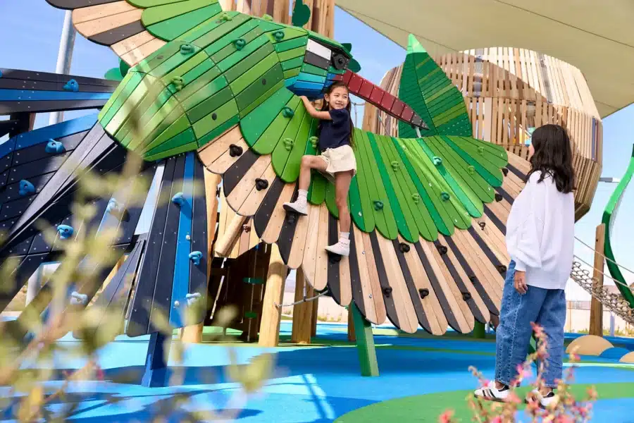 A child climbs a large, bird-shaped playground structure while an adult stands nearby watching. The scene is bright and outdoors with a blue play surface.