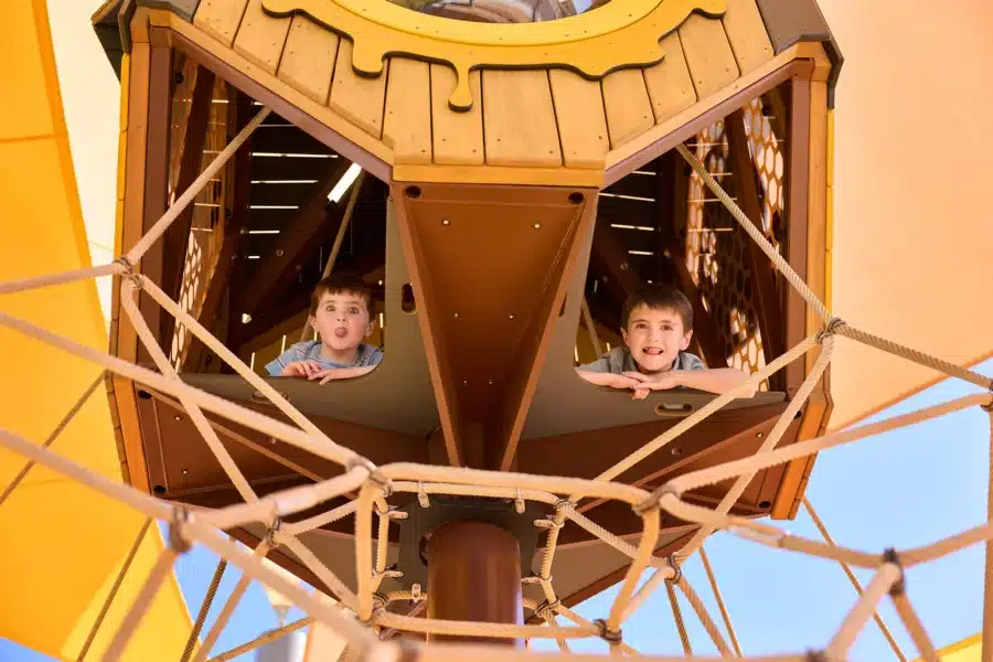 Two children look out from a wooden play structure, surrounded by ropes and shaded by a yellow canopy, with blue sky visible in the background.