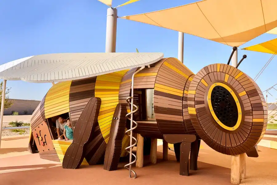 Large playground structure shaped like a bee with children playing inside, featuring wood paneling, slides, and a climbing spiral under sunshades.