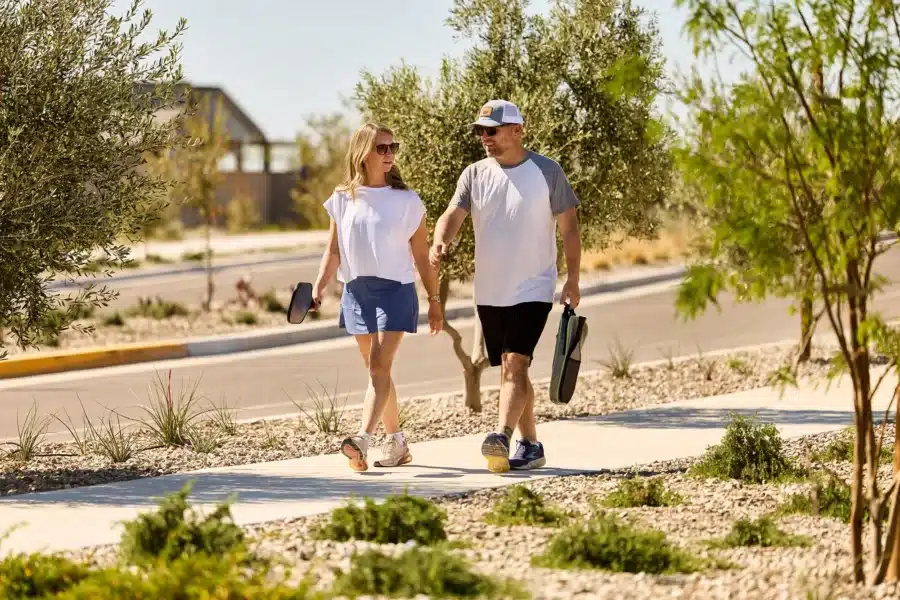 Two people walk on a sunny sidewalk, each holding a paddle, surrounded by trees and plants, with a road visible beside them.