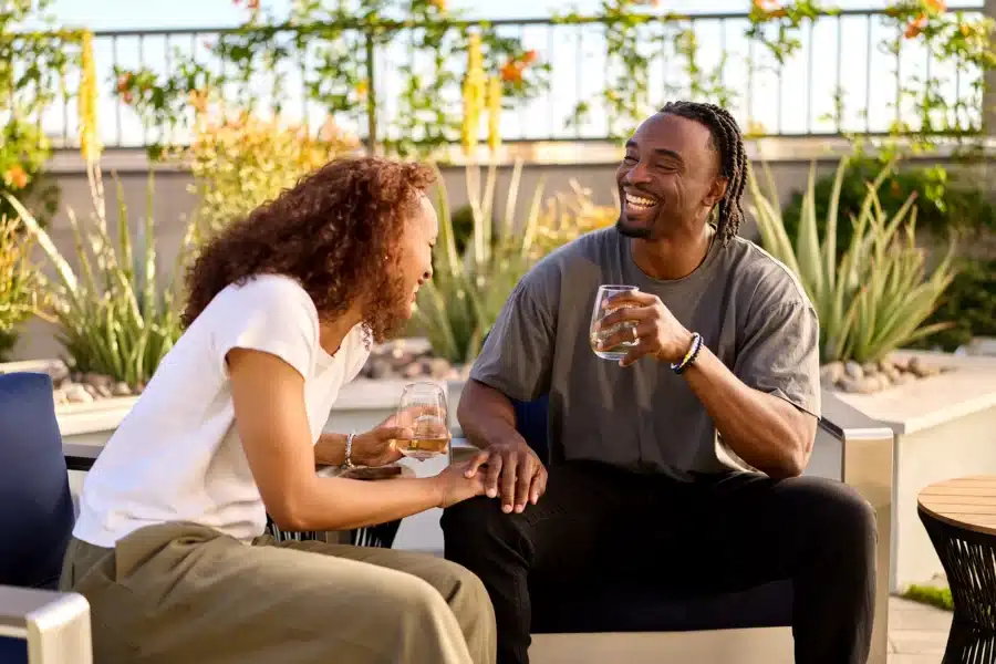 Two people sit outdoors on a patio, smiling and laughing while holding drinks. Plants and a fence are visible in the background.
