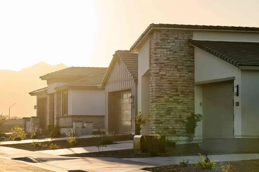 Row of modern single-story suburban homes with stone and stucco exteriors, garage doors, and landscaped front yards in sunlight.